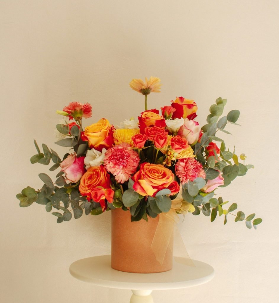 Arreglo floral Sunny de Celiflor con rosas amarillas, naranjas y rosadas, lisianthus, solidago y follajes naturales en base color salmón.