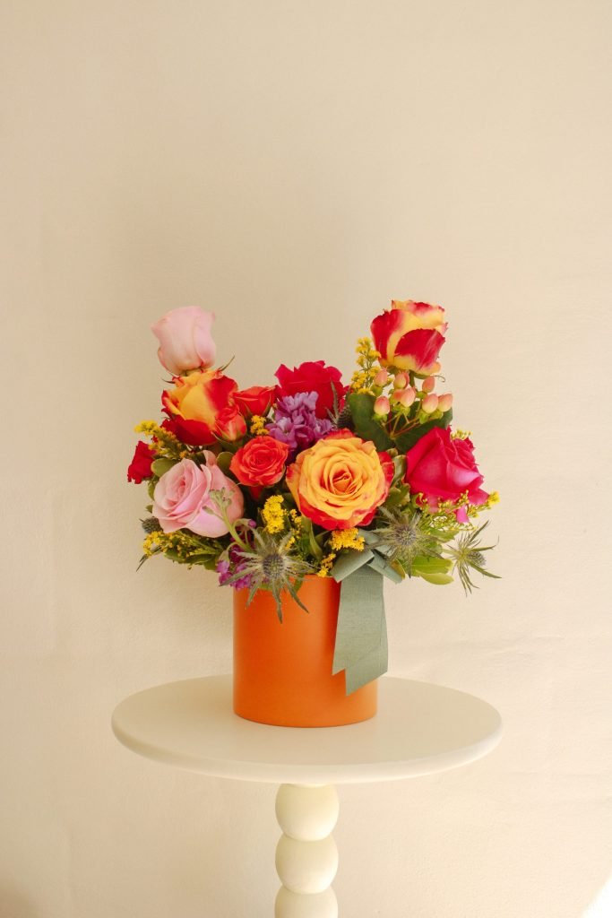 Arreglo Viva de Celiflor con rosas fucsias, rosadas y naranjas, mini rosas, solidago y stock en base color naranja, creación artesanal ideal para regalar o decorar.